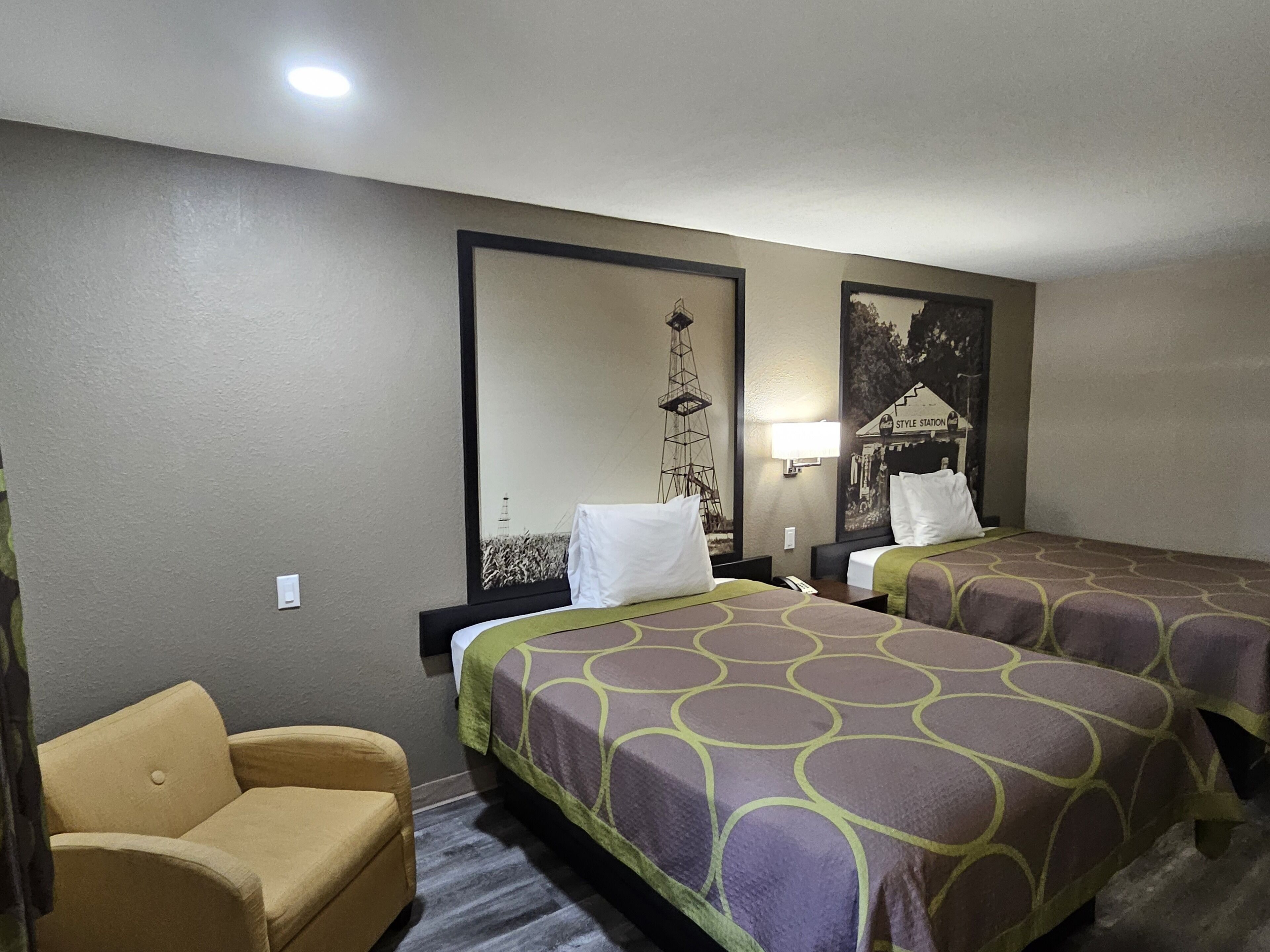 A hotel room with two beds, a chair, a lamp, and a wall mural.