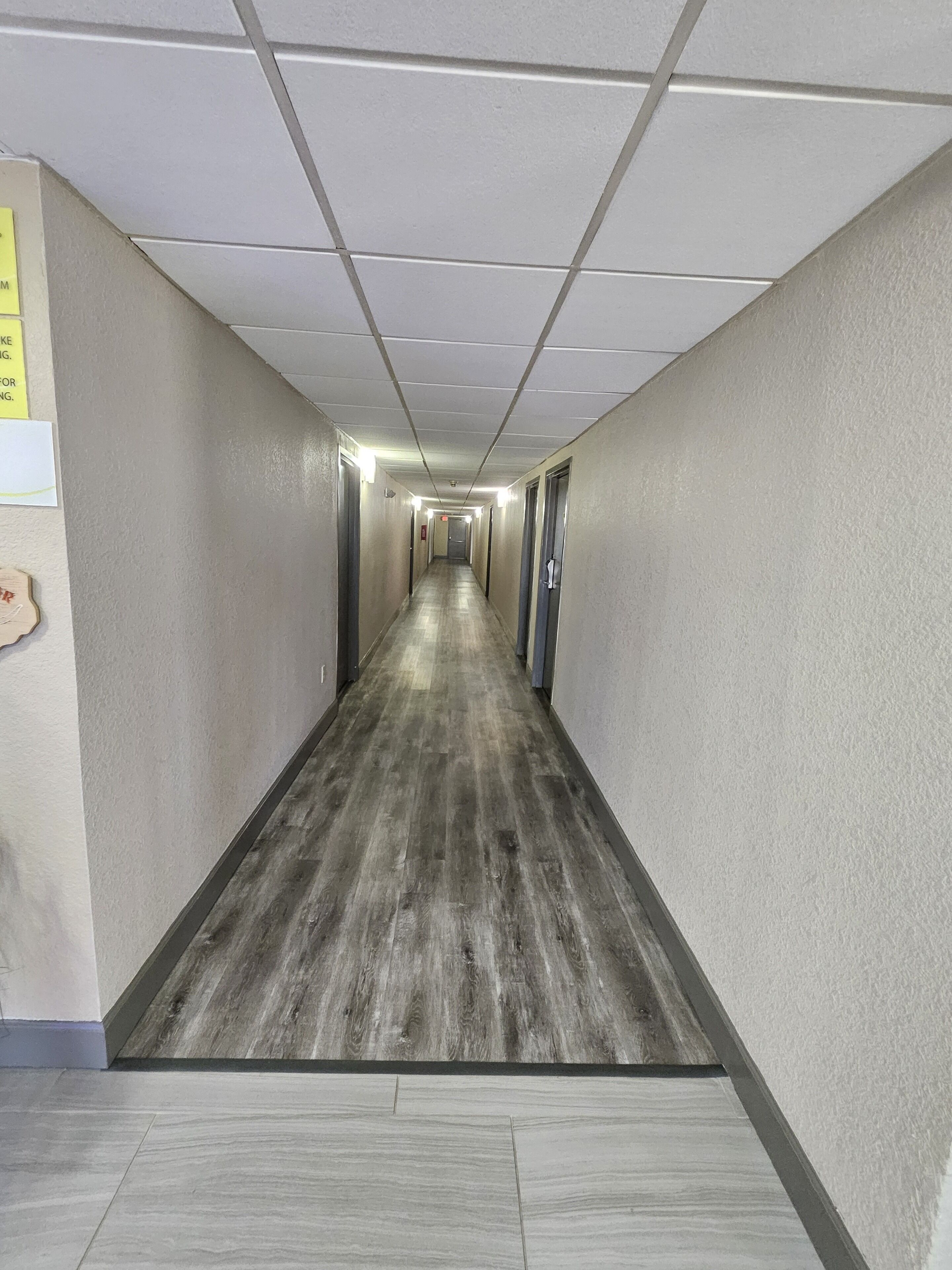 Um corredor comprido e estreito com piso de madeira e paredes brancas. Há portas em ambos os lados e uma placa na parede esquerda.
