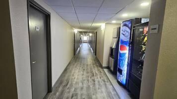 Un couloir d’hôtel avec un plancher en bois, des portes des deux côtés et des distributeurs automatiques sur la droite.