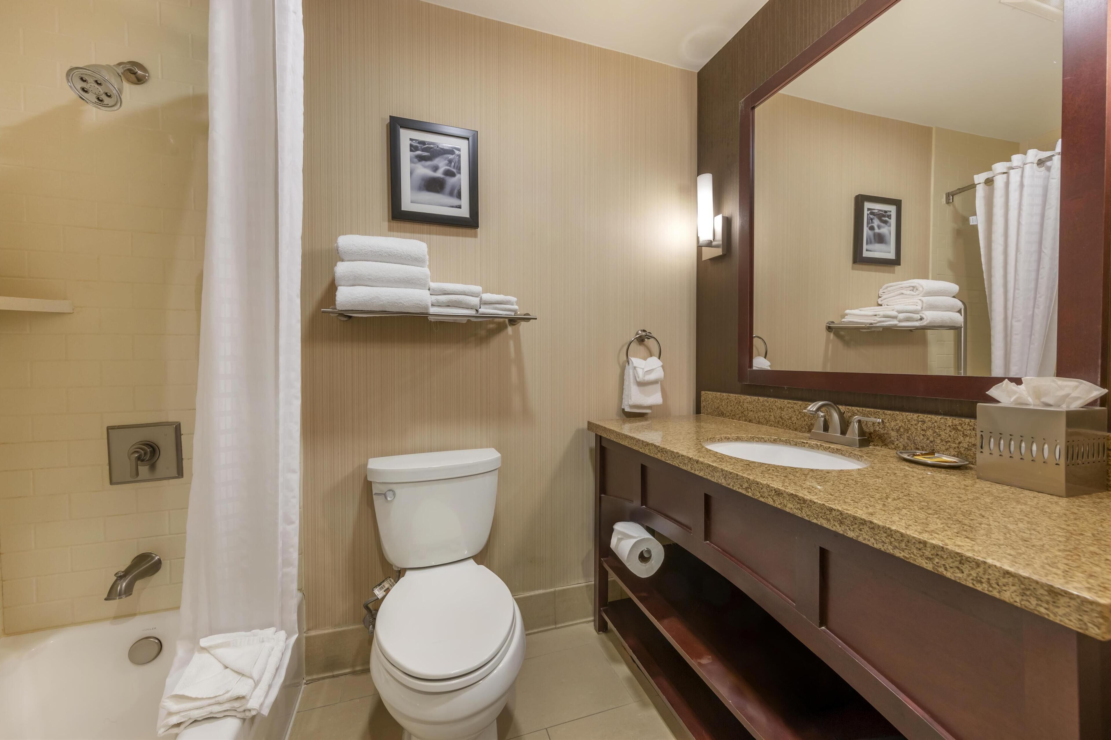 A bathroom with a toilet, sink, and shower. There are towels on a shelf and a mirror above the sink.