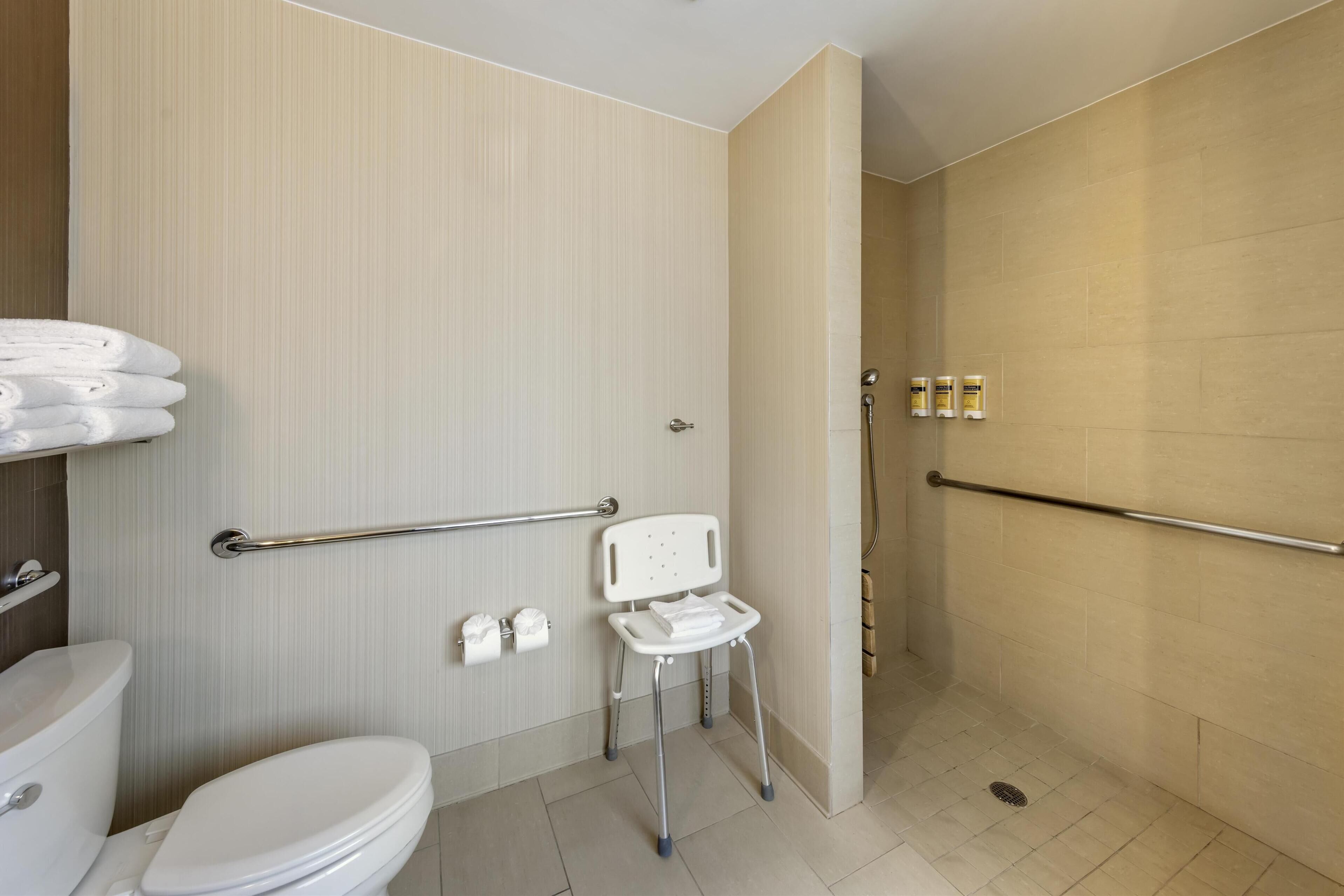 Une salle de bain moderne équipée de toilettes, d’une barre d’appui, d’un espace douche et d’un banc accessible aux personnes en fauteuil roulant.