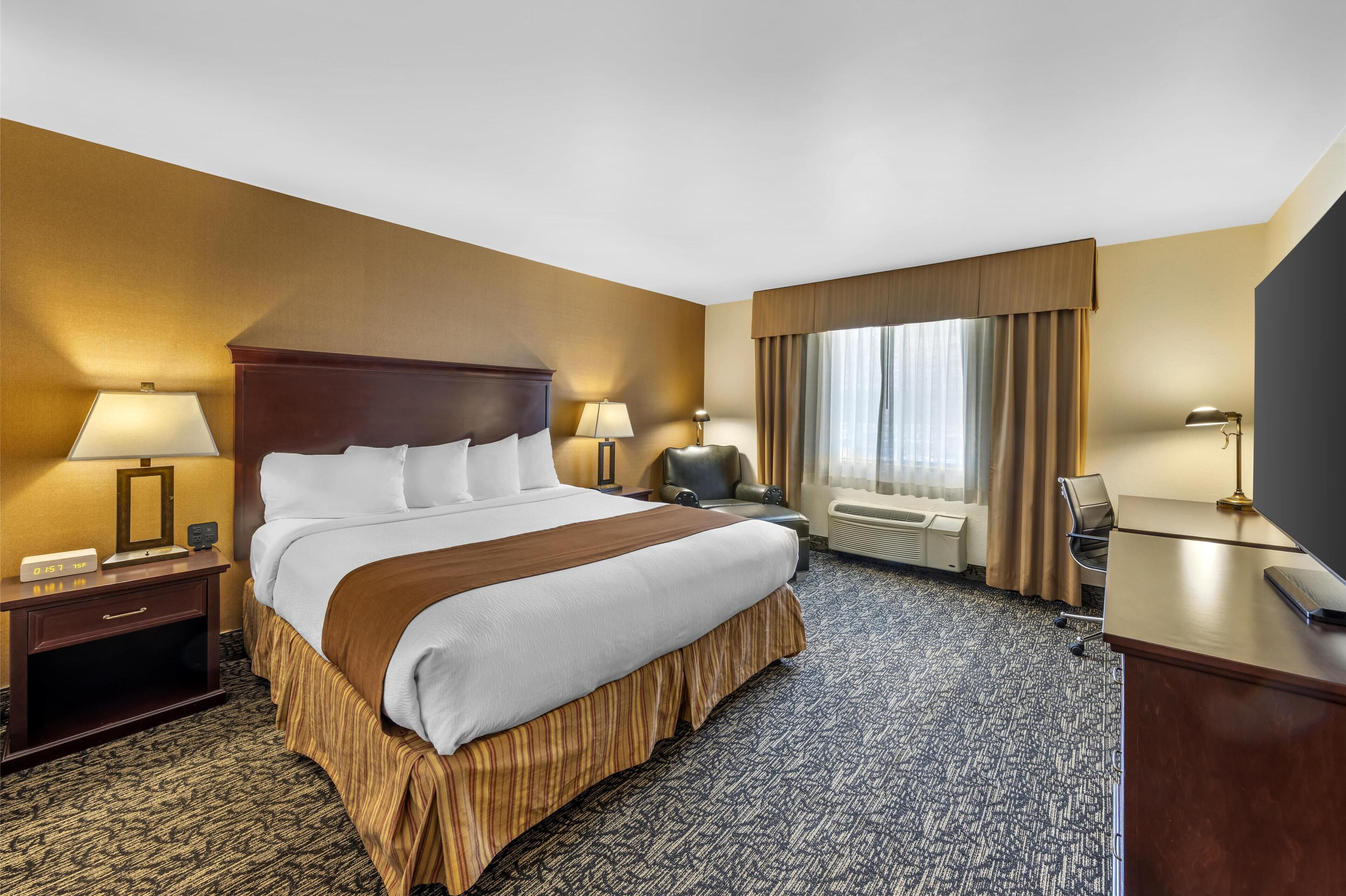 Habitación de hotel con una cama grande, un escritorio con computadora, una silla, un ventanal con cortinas y un reloj de pared.