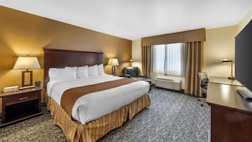 Habitación de hotel con una cama grande, un escritorio con computadora, una silla, un ventanal con cortinas y un reloj de pared.