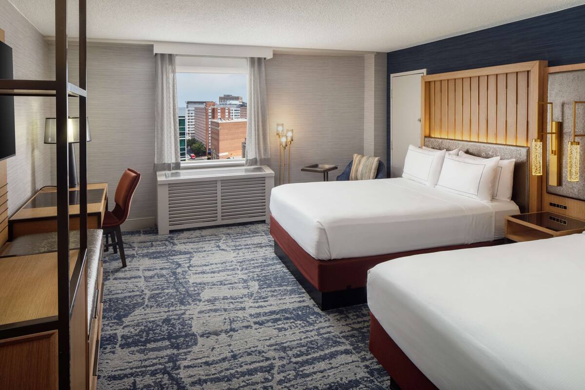 A hotel room with a large bed, a desk, a chair, a small table, and a window with a city view.