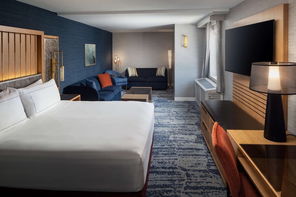 A hotel room with a large bed, a sofa, a desk with a lamp, a TV, and a wall-mounted artwork.