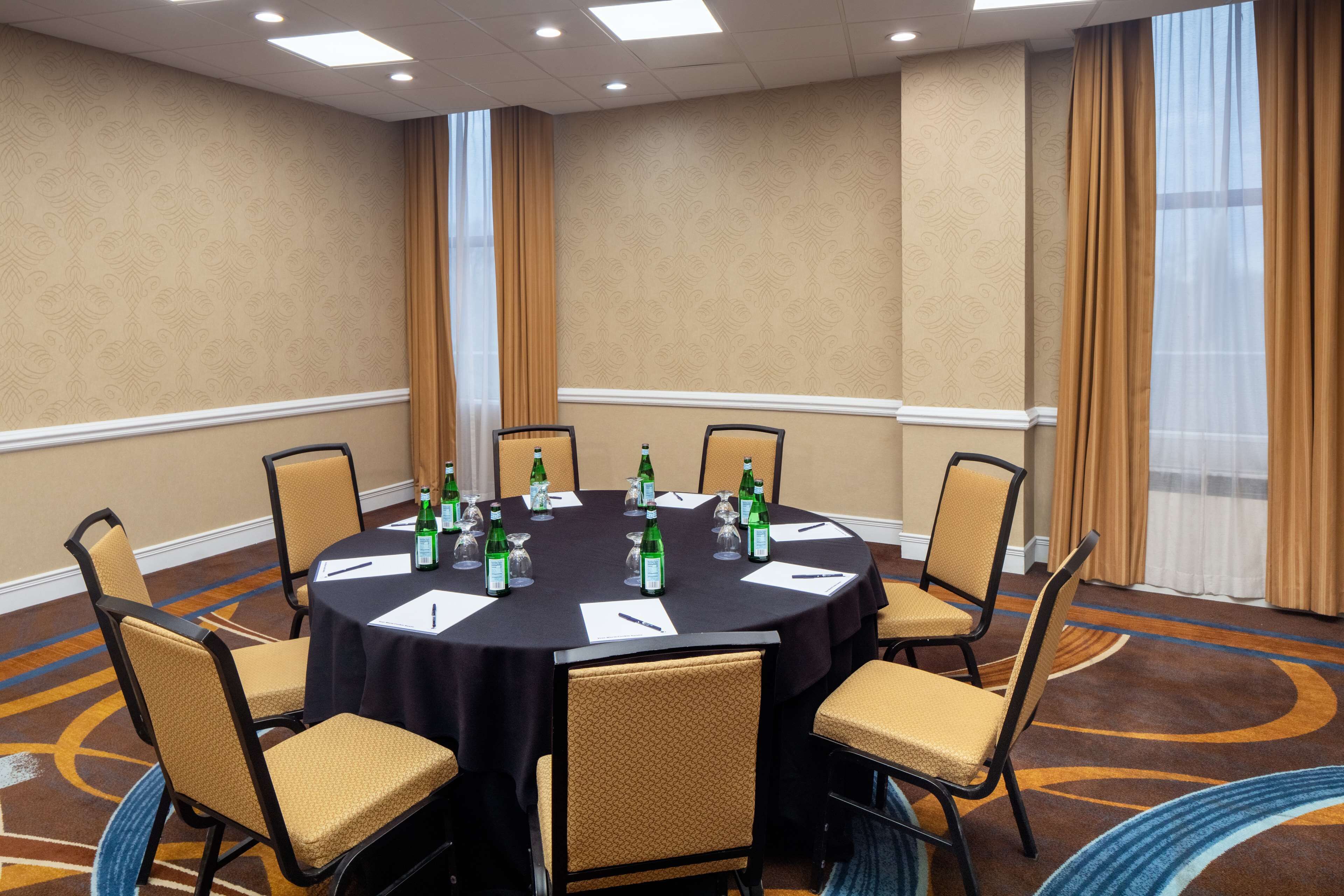 A conference room with a round table, six chairs, and a black tablecloth. There are bottles and glasses on the table, and the room has beige walls and a carpeted floor.