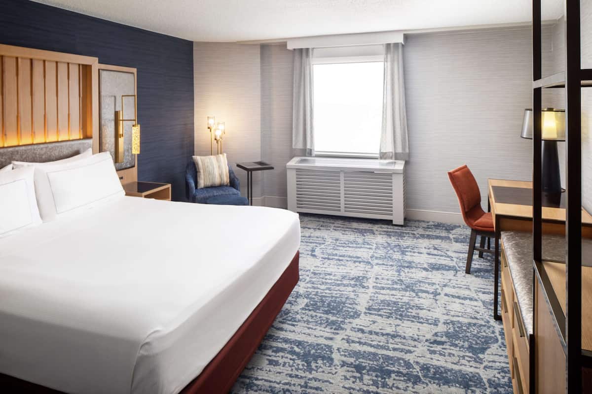 A hotel room with a large bed, a desk with a chair, a small table, a lamp, and a window with curtains.