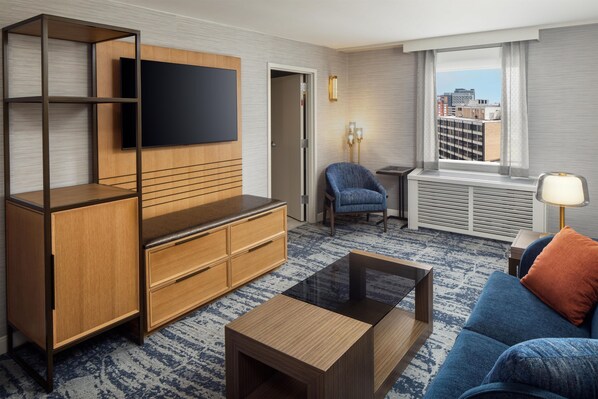 A modern hotel room with a flat-screen TV mounted on a wooden cabinet, a sofa, a coffee table, and a view of the city through a large window.