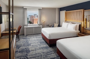 A hotel room with a bed, a desk, a chair, a window with a view of buildings, and a wall-mounted light fixture.