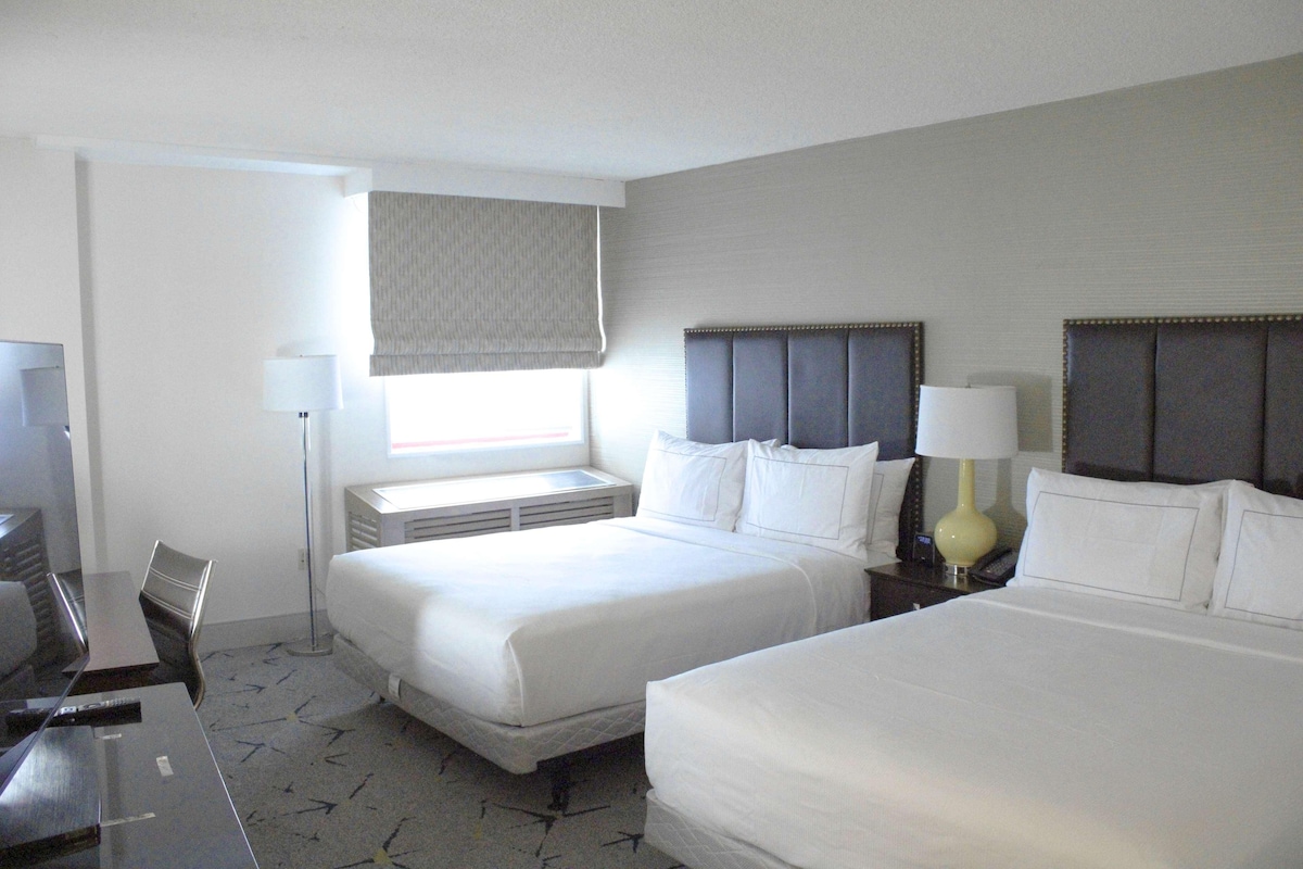 A hotel room with two beds, a desk, a chair, a lamp, and a window with a curtain.