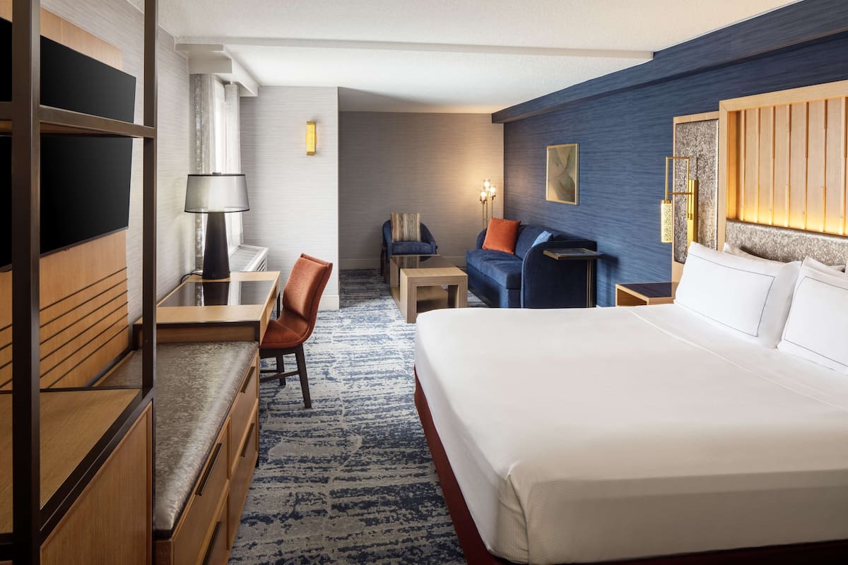 A hotel room with a large bed, a desk with a lamp, a chair, a sofa, and a small table.