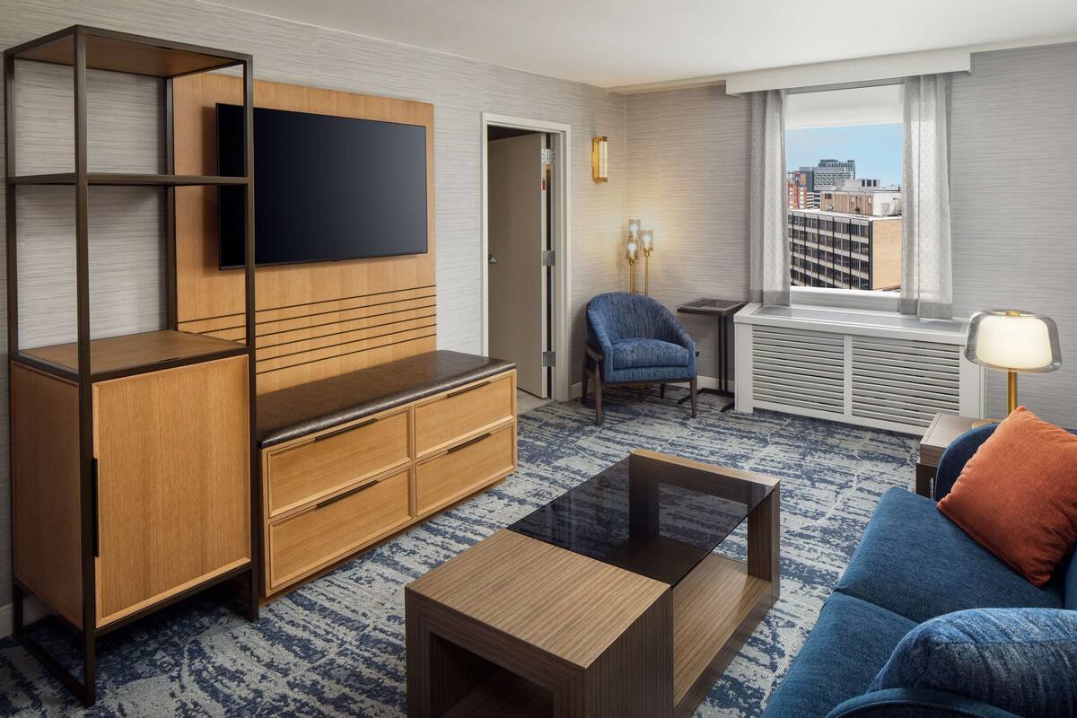 A modern hotel room with a flat-screen TV mounted on the wall, a wooden entertainment unit, a blue sofa with an orange pillow, a coffee table, and a view of the city through a window.
