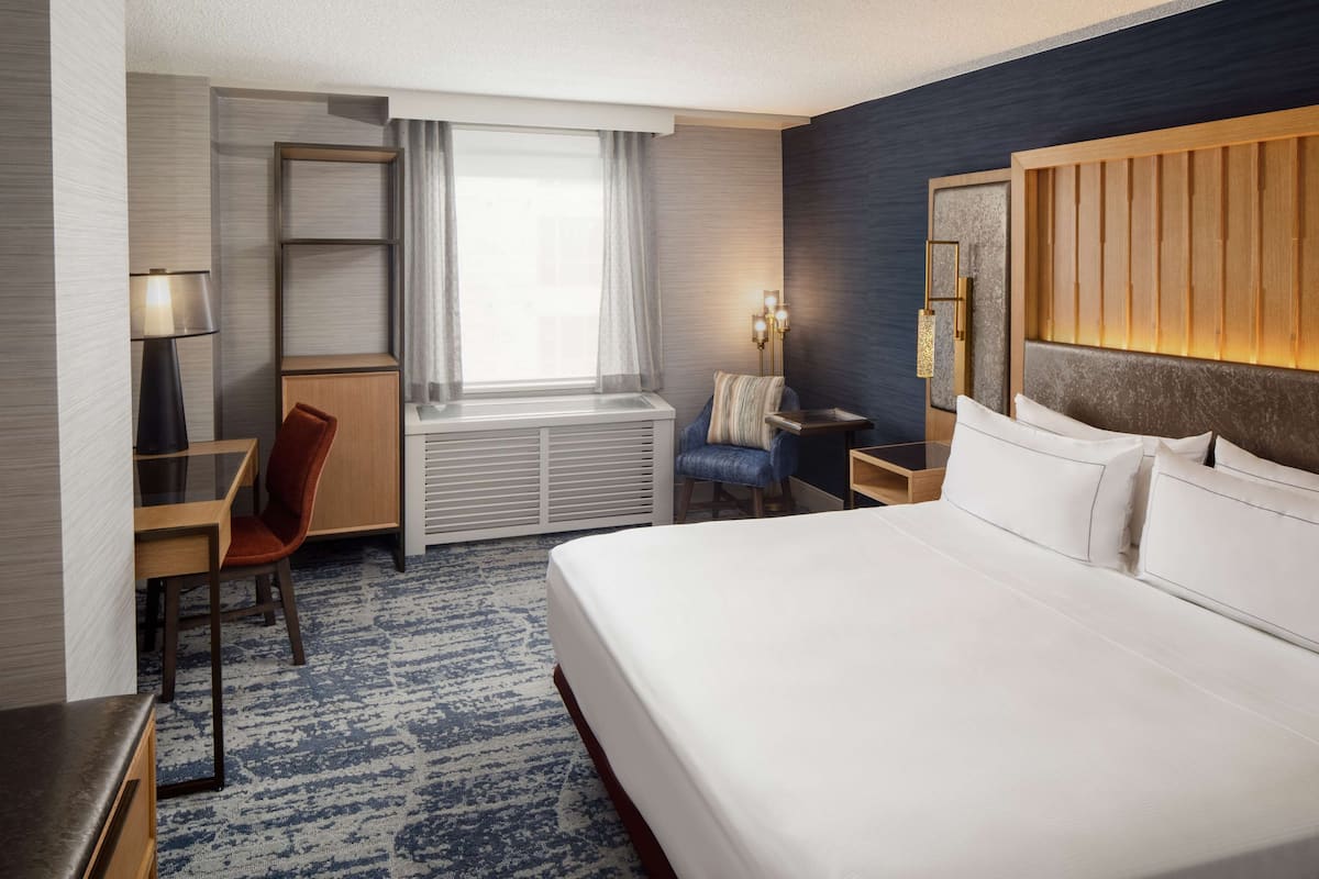 A hotel room with a large bed, a desk with a chair, a small table, a lamp, a window with curtains, and a headboard with a light panel.