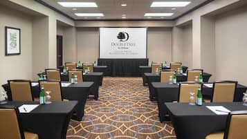 Uma sala de conferências com mesas e cadeiras, uma tela de projetor exibindo o logotipo da DoubleTree by Hilton, e piso coberto de carpete.