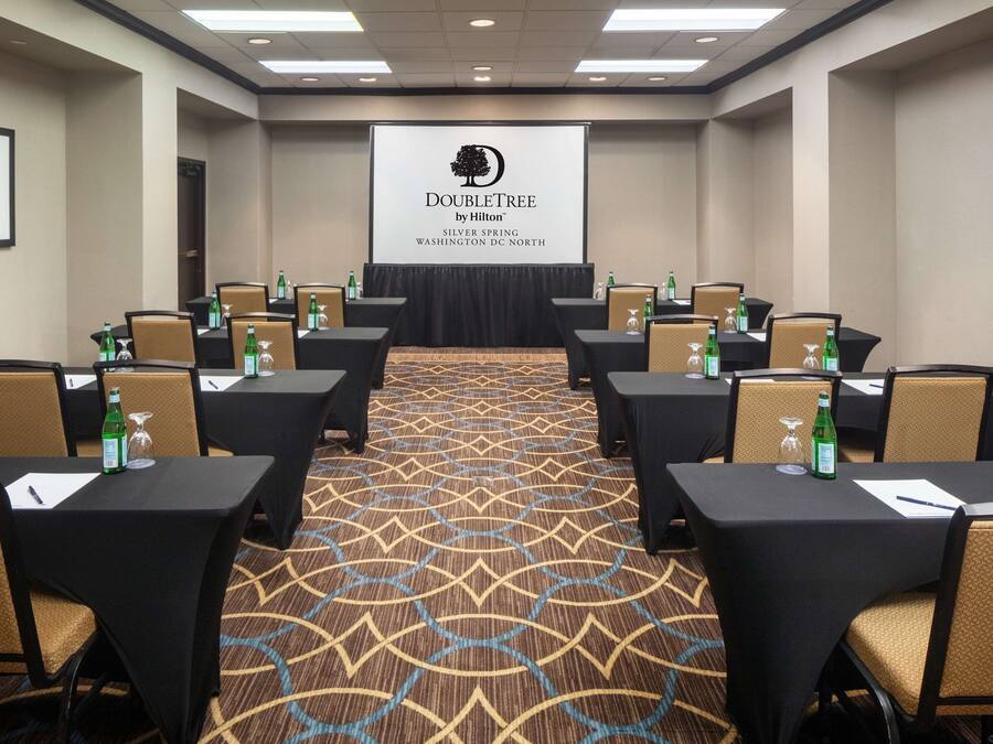 Uma sala de conferências com mesas e cadeiras, uma tela de projetor exibindo o logotipo da DoubleTree by Hilton, e piso coberto de carpete.