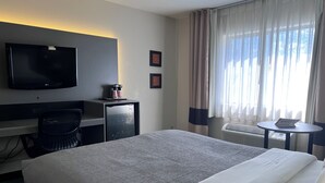 Habitación de hotel con cama, escritorio con televisor, cafetera, silla, mesita con revista y ventana con cortinas.