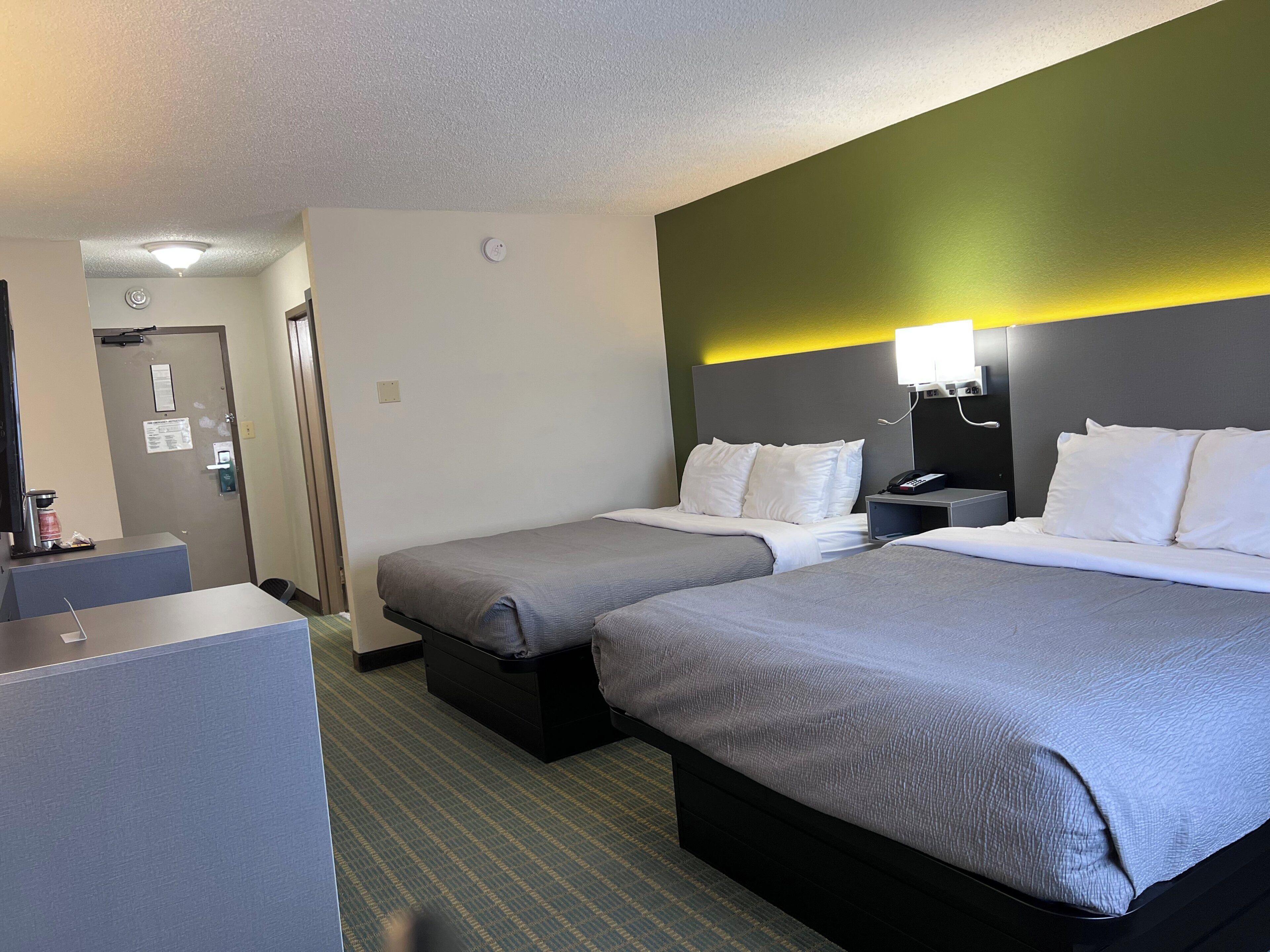 A hotel room with two beds, a desk, a coffee machine, and a door with a sign.