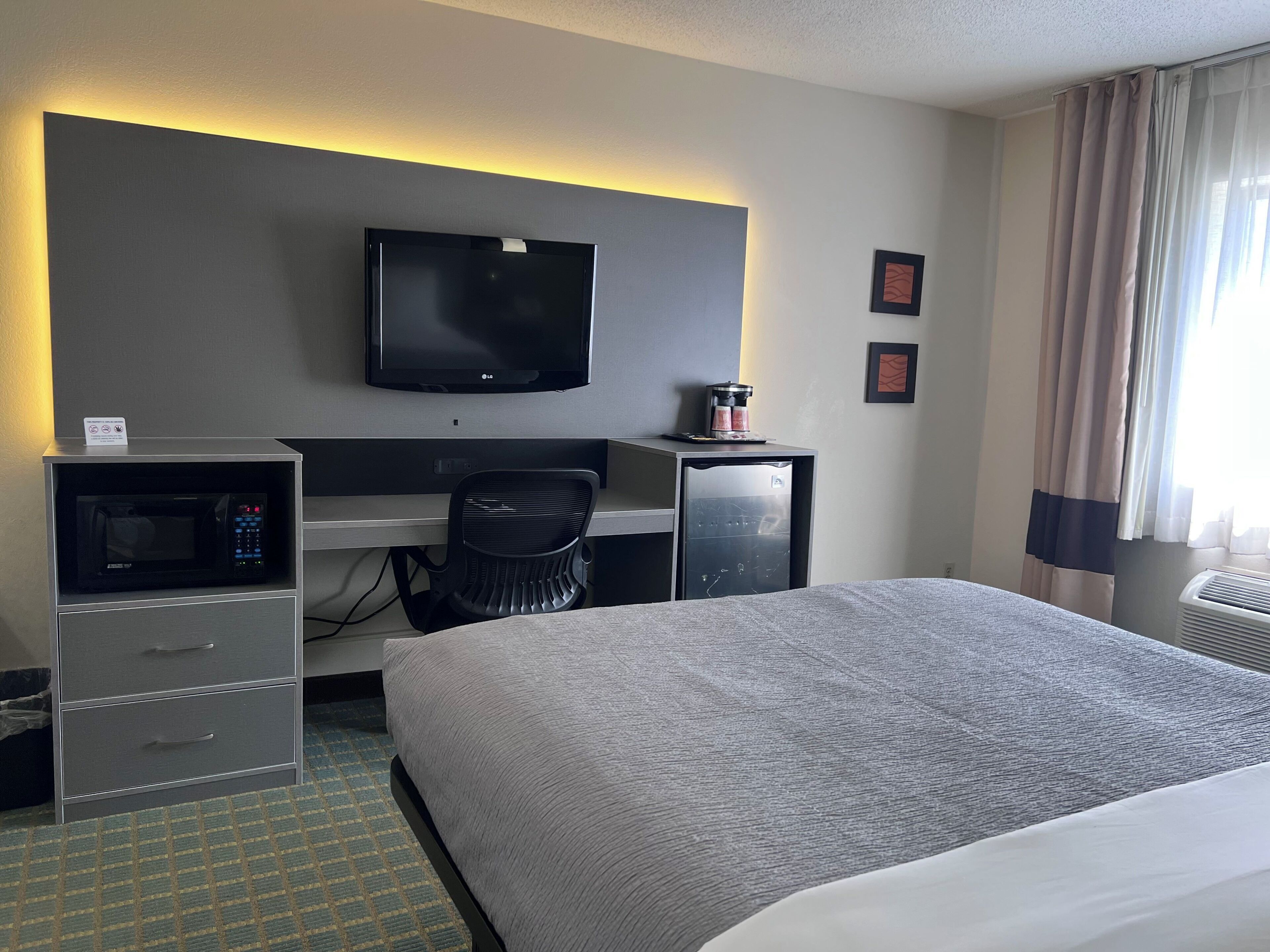 Et hotelværelse med en seng, et skrivebord med en computer, en mikroovn, en stol og et fjernsyn.