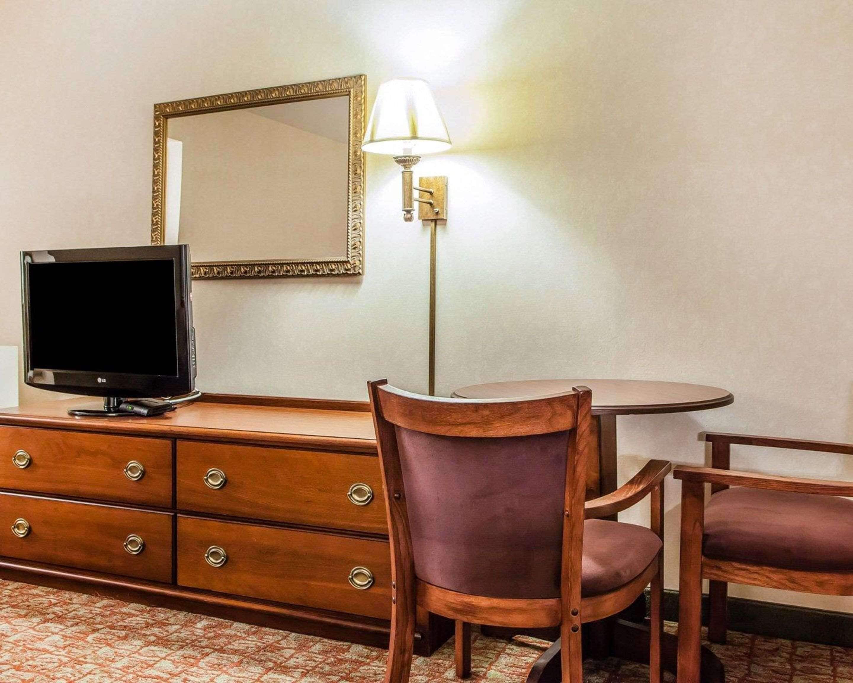 Ein Hotelzimmer mit einem Holzkoffer, einem Fernseher, einem runden Tisch, zwei Stühlen, einem Spiegel und einer Wandlampe.