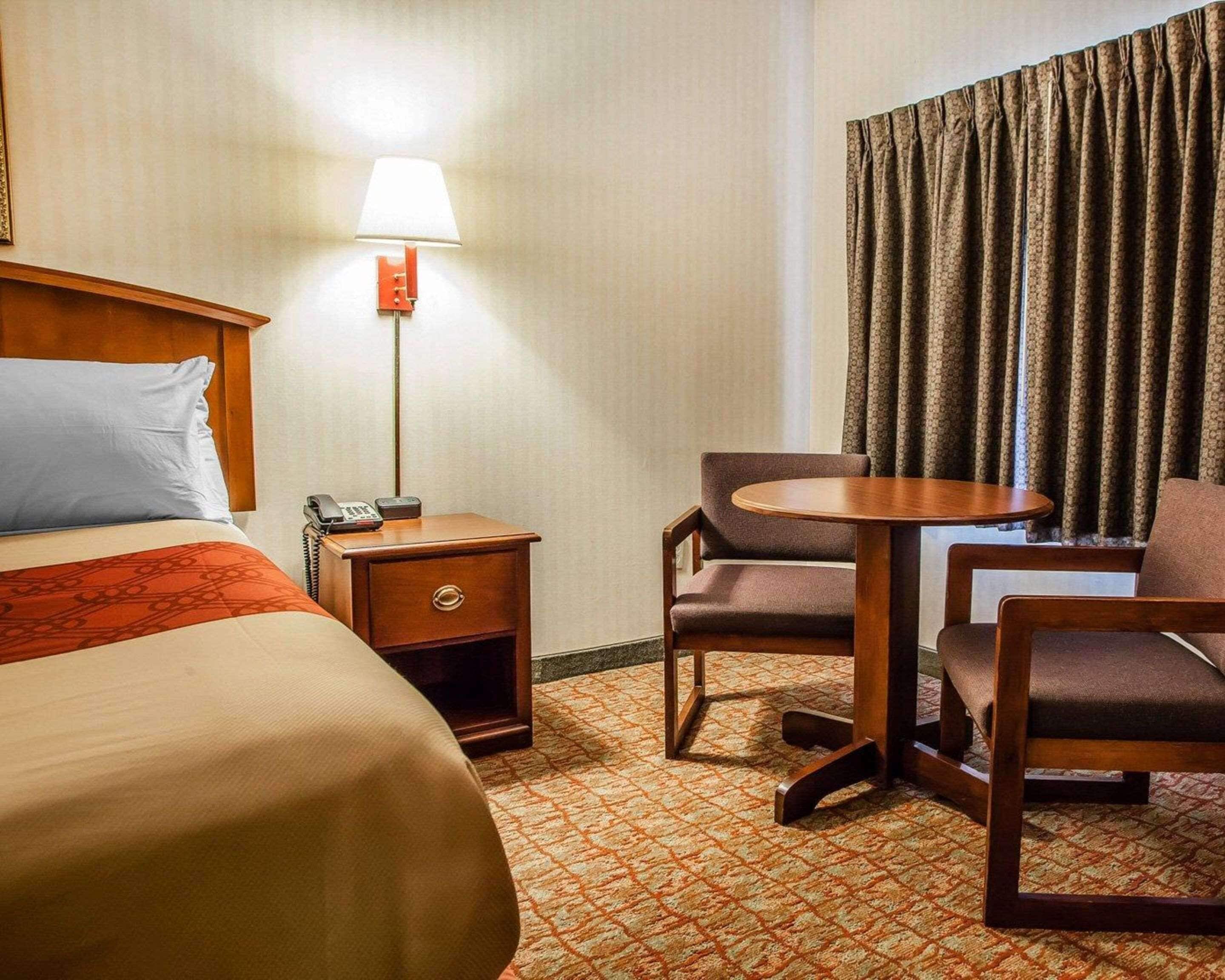 A hotel room with a bed, bedside table with a lamp, a small table with chairs, and a window with curtains.