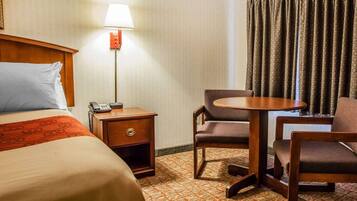 A hotel room with a bed, bedside table with a lamp, a small table with chairs, and a window with curtains.