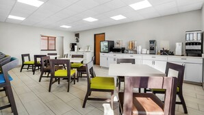 Una cocina moderna con armarios blancos, microondas, cafetera y refrigerador. Hay sillas de madera con cojines verdes y una mesa blanca.