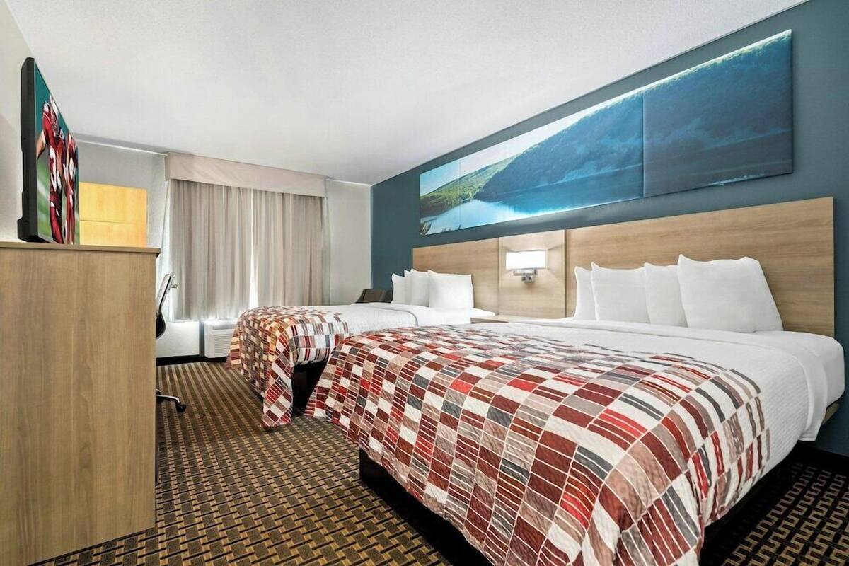 Une chambre d’hôtel avec deux lits, une grande fenêtre avec des rideaux, un tableau au-dessus des lits et une tête de lit en bois.