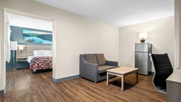 A hotel room with a bed, a sofa, a table, a chair, a desk, a lamp, and a refrigerator.