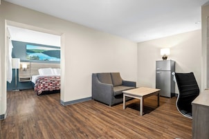 Quarto de hotel com cama, sofá, mesa, cadeira, escrivaninha, luminária e geladeira.