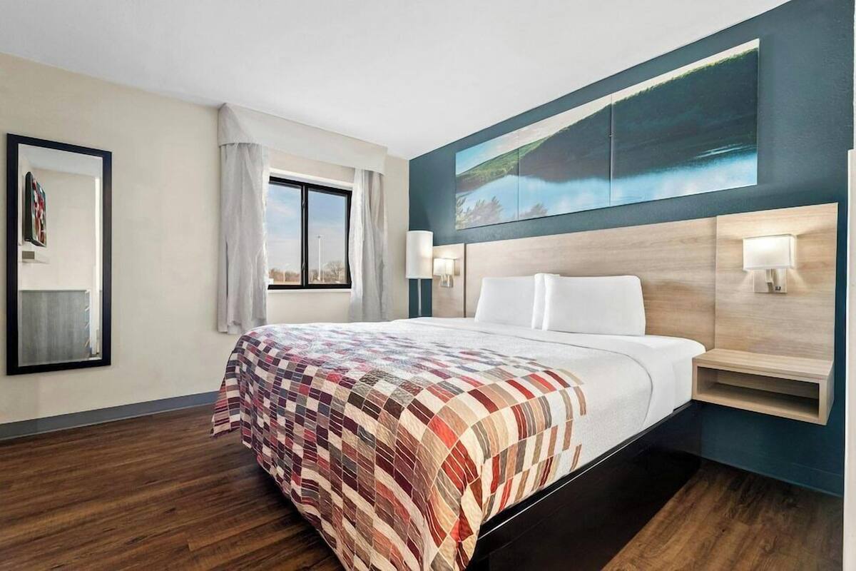 Une chambre d’hôtel avec un grand lit, une fenêtre avec des rideaux, un tableau au mur et une table de chevet avec une lampe.