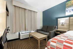 A hotel room with a bed, a small table, a sofa, a window with curtains, a wall-mounted lamp, and a radiator. - Red Roof Inn & Suites Tomah (Tomah)