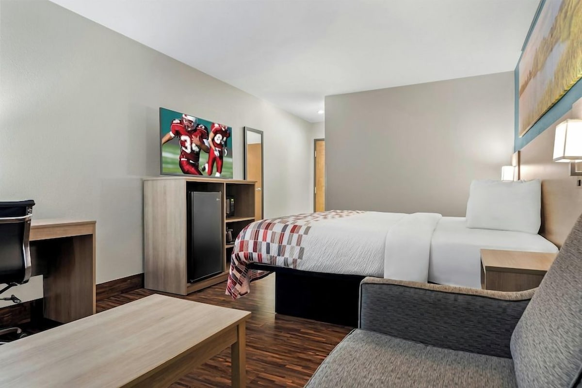 A hotel room with a bed, a desk, a chair, a TV, and a painting on the wall.