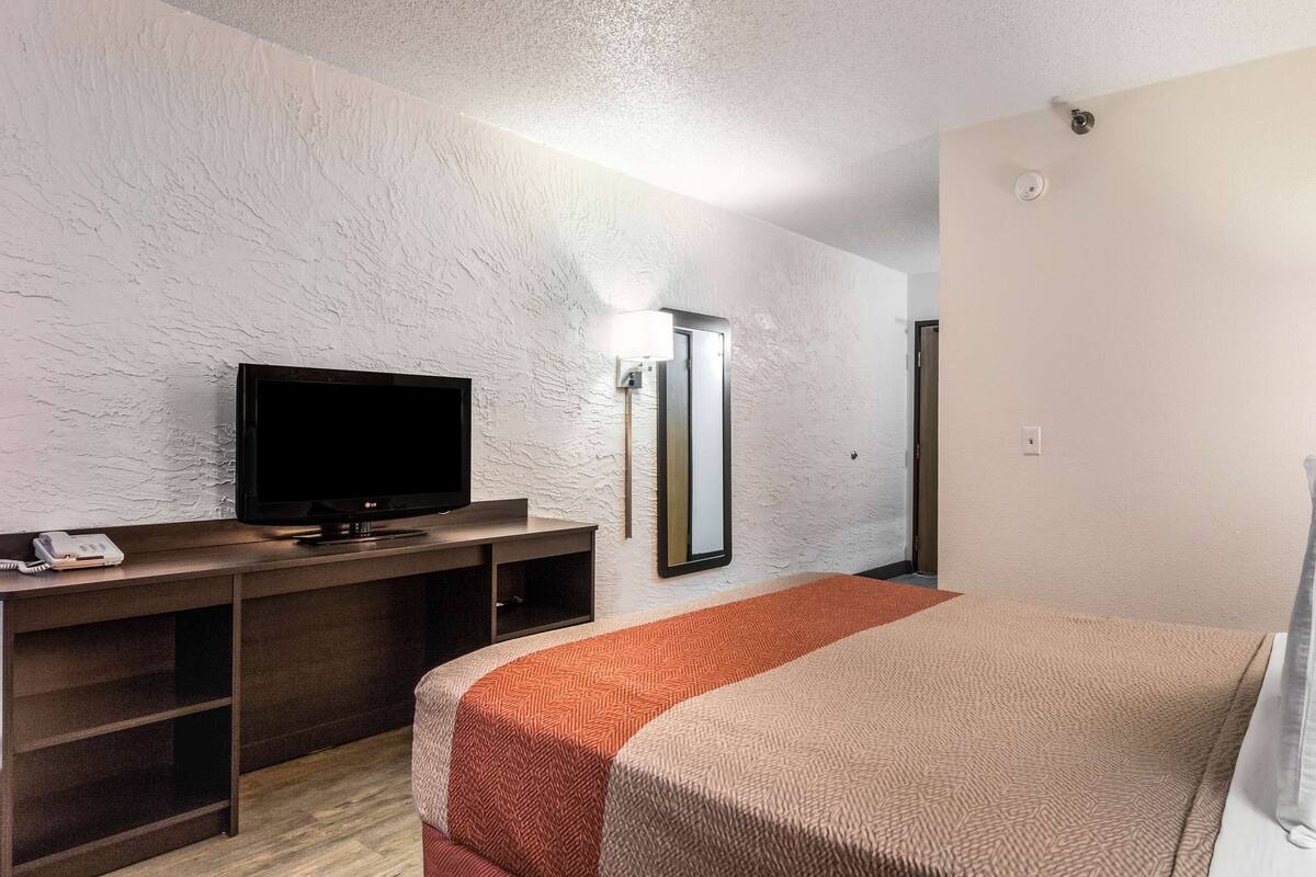 A hotel room with a bed, a TV on a wooden stand, a mirror, and a lamp.