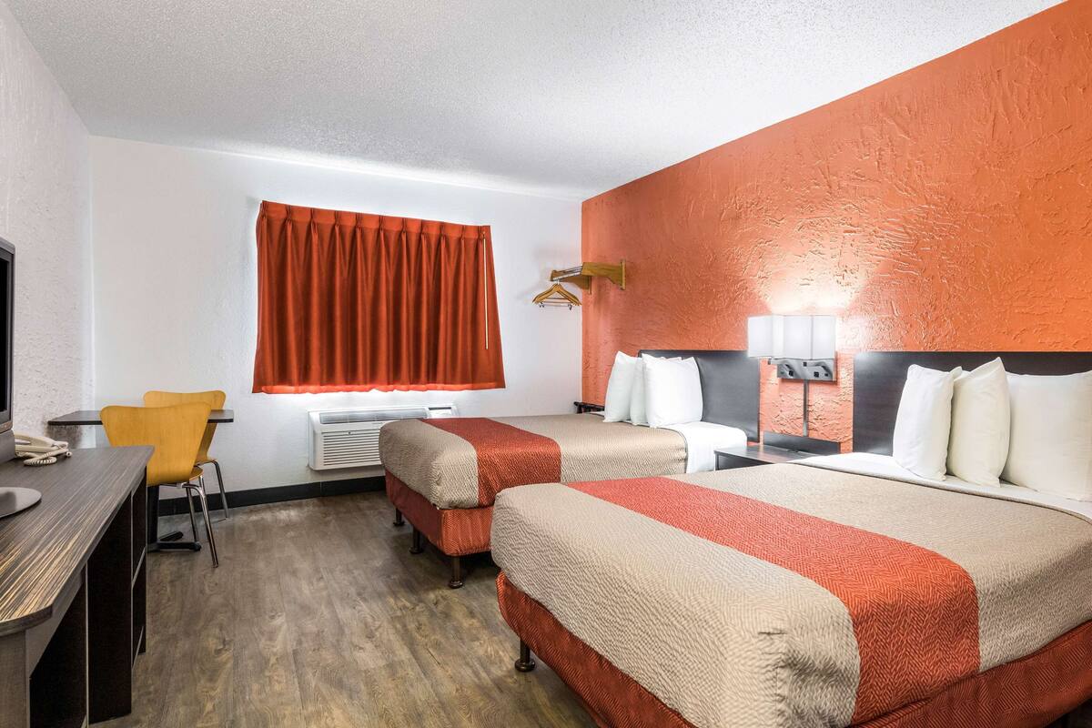 A hotel room with two beds, a desk, a chair, and a window with a red curtain.