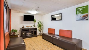 A waiting area with two brown sofas, a flat-screen TV mounted on the wall, a fireplace, and a green poster advertising an app. - Motel 6 Elk Grove Village, IL (Elk Grove Village)