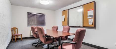 A conference room with a wooden table, six pink upholstered chairs, a wooden chair, a window with blinds, and two bulletin boards on the wall.
