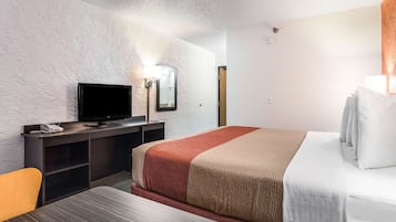 A hotel room with a bed, a desk with a television, a mirror, and a bedside table.