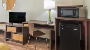 Habitación de hotel con escritorio, silla, televisor, lámpara, microondas y cubo de basura.