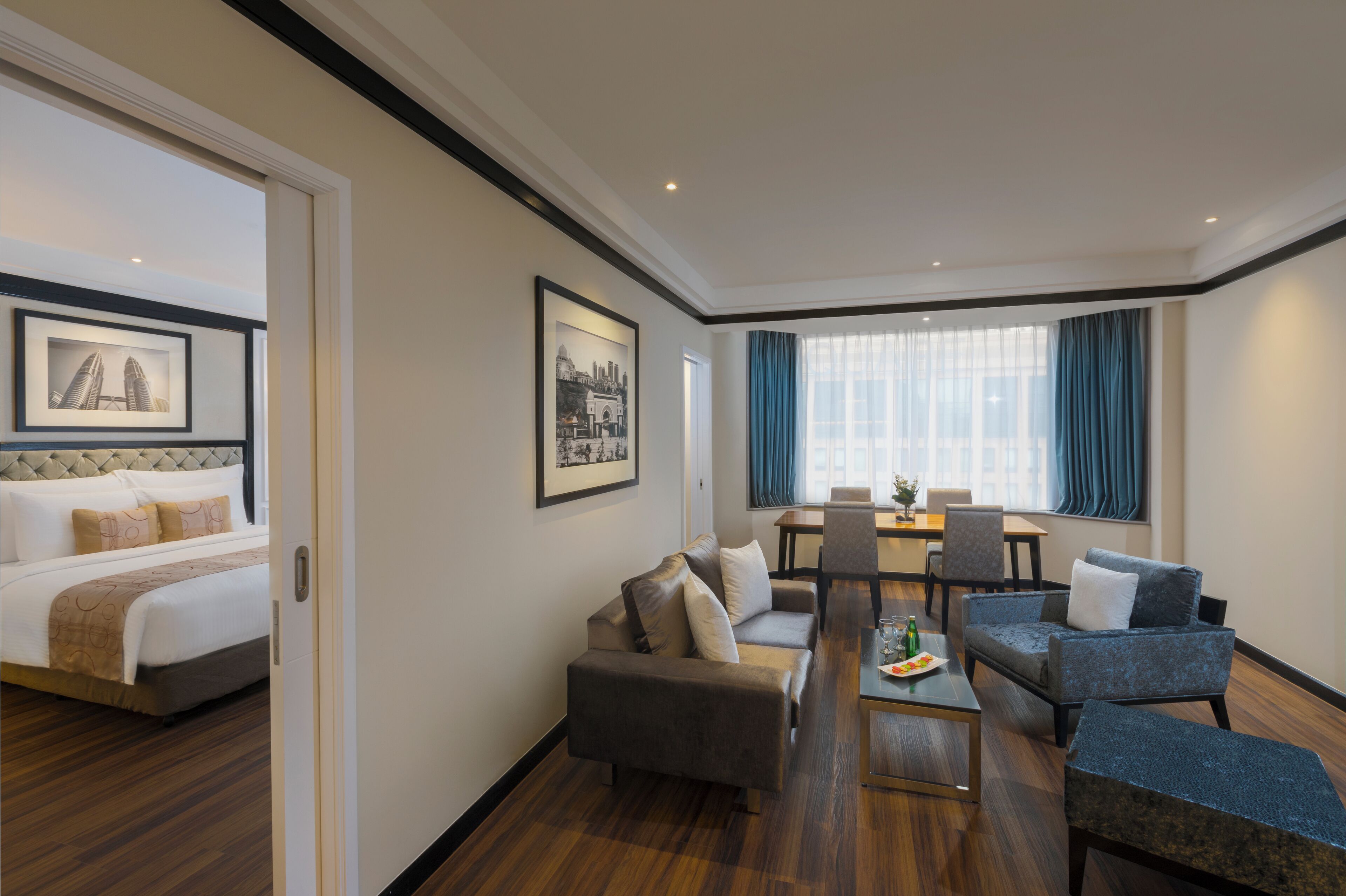 A hotel room with a bed, a sofa, a dining table with chairs, and a view of the city through large windows.