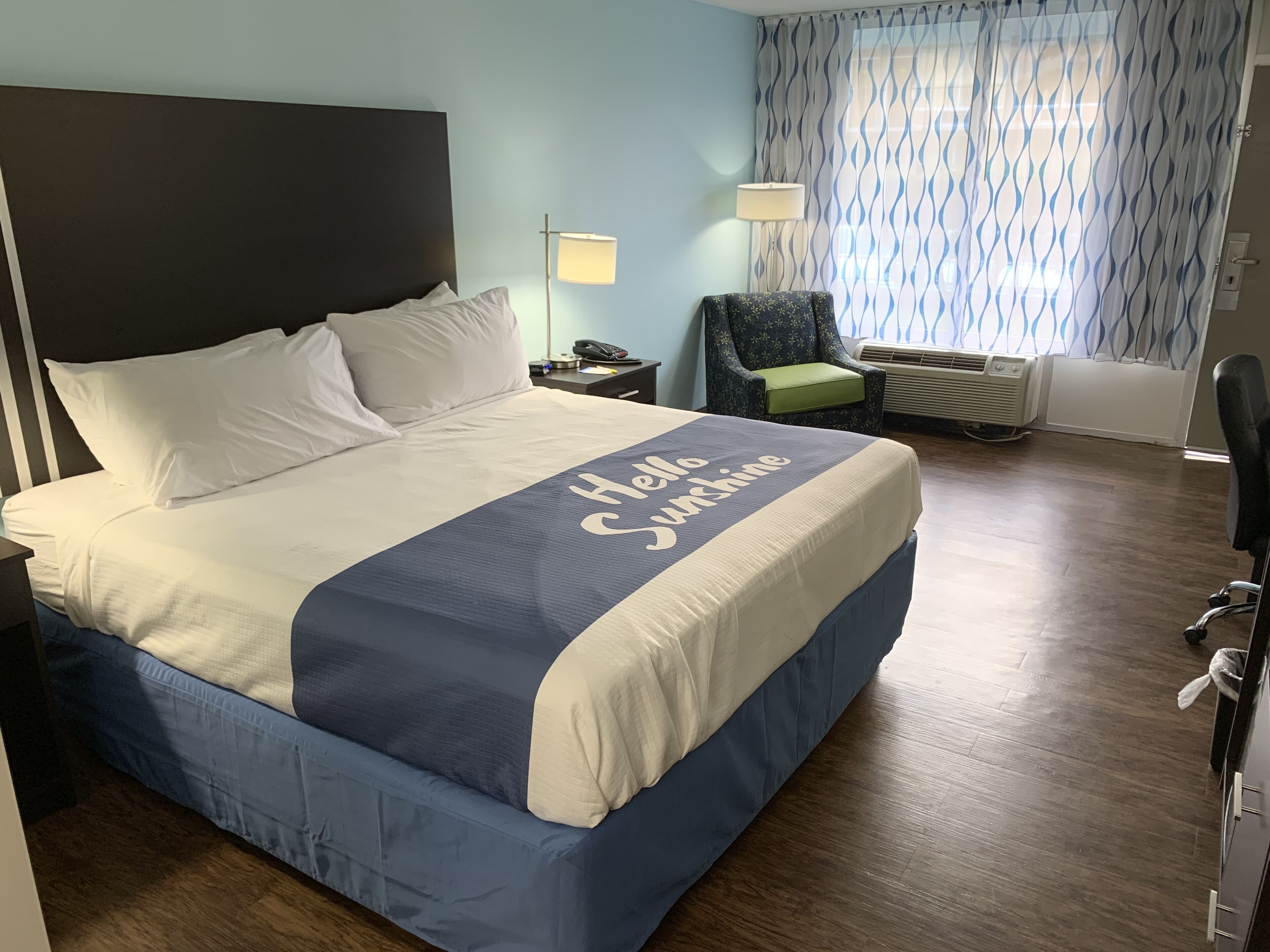 A hotel room with a bed, a nightstand, a chair, a lamp, and an air conditioning unit.