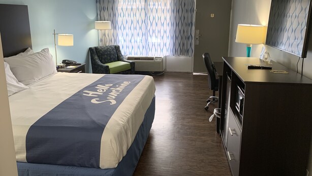 A hotel room with a bed, a desk, a chair, a desk lamp, a wall-mounted TV, and a window with patterned curtains.