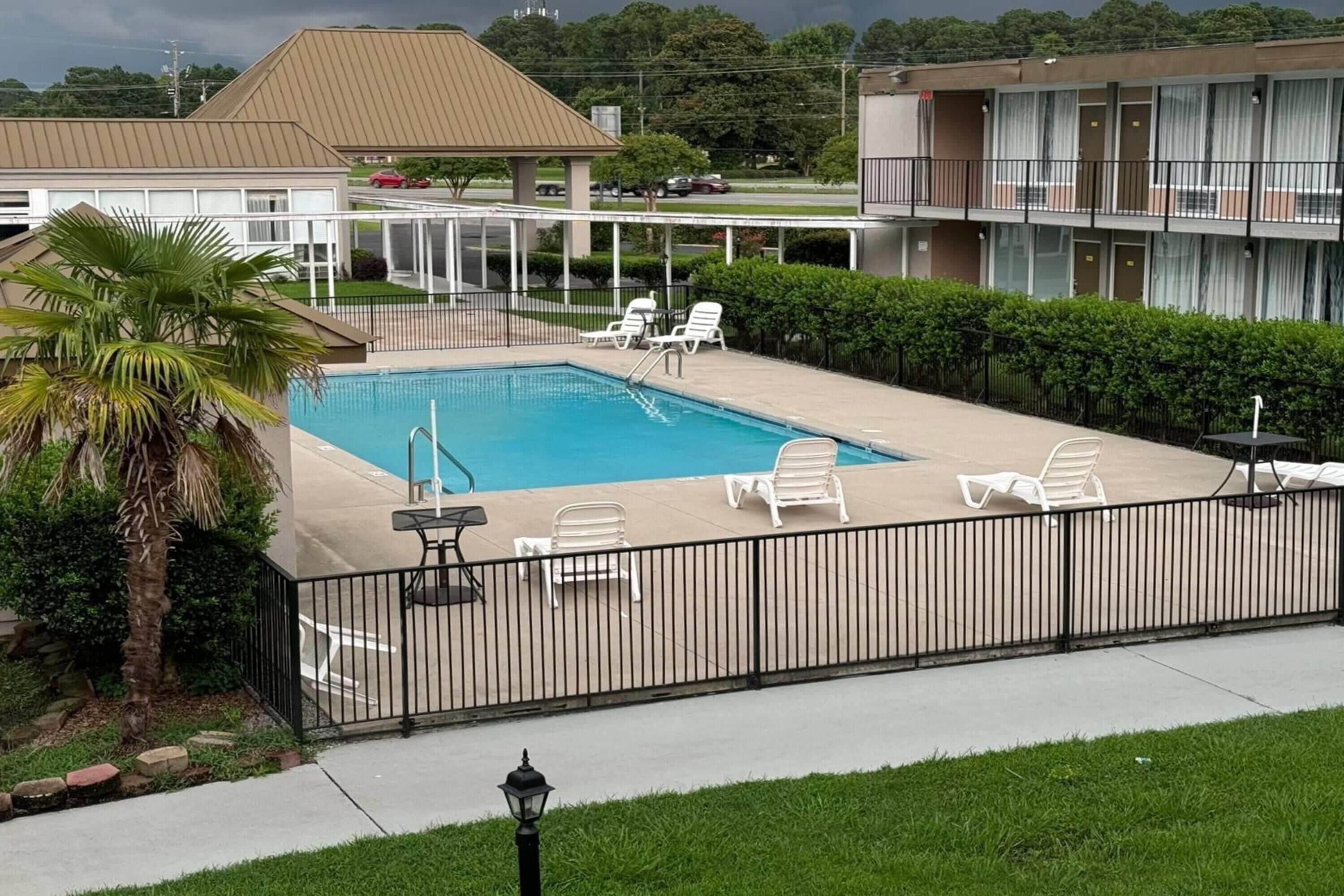 Un área de piscina con sillones de descanso, una palmera y un edificio con balcón al fondo.