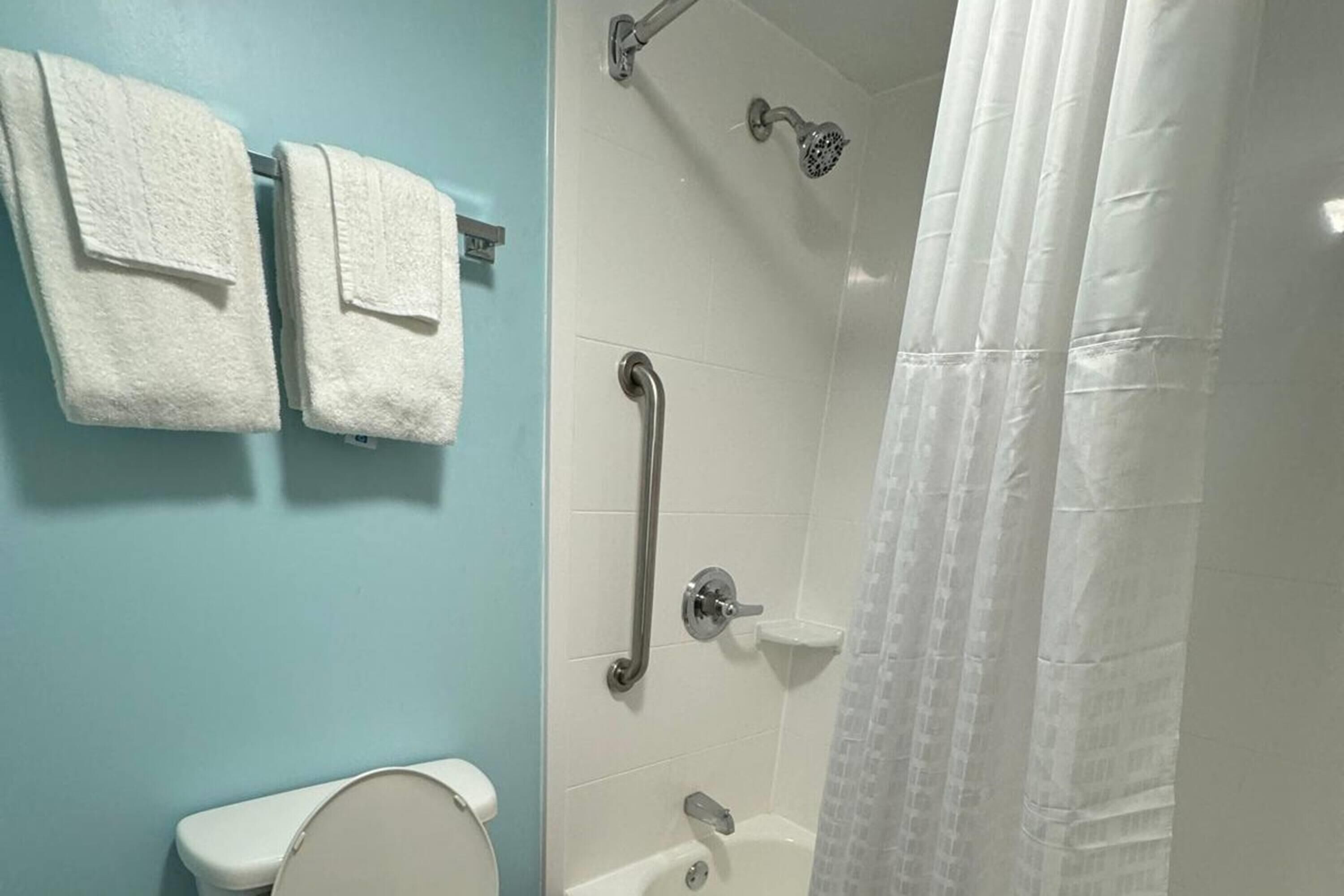 Un baño con inodoro, ducha con barra de apoyo y dos toallas colgadas en una pared.