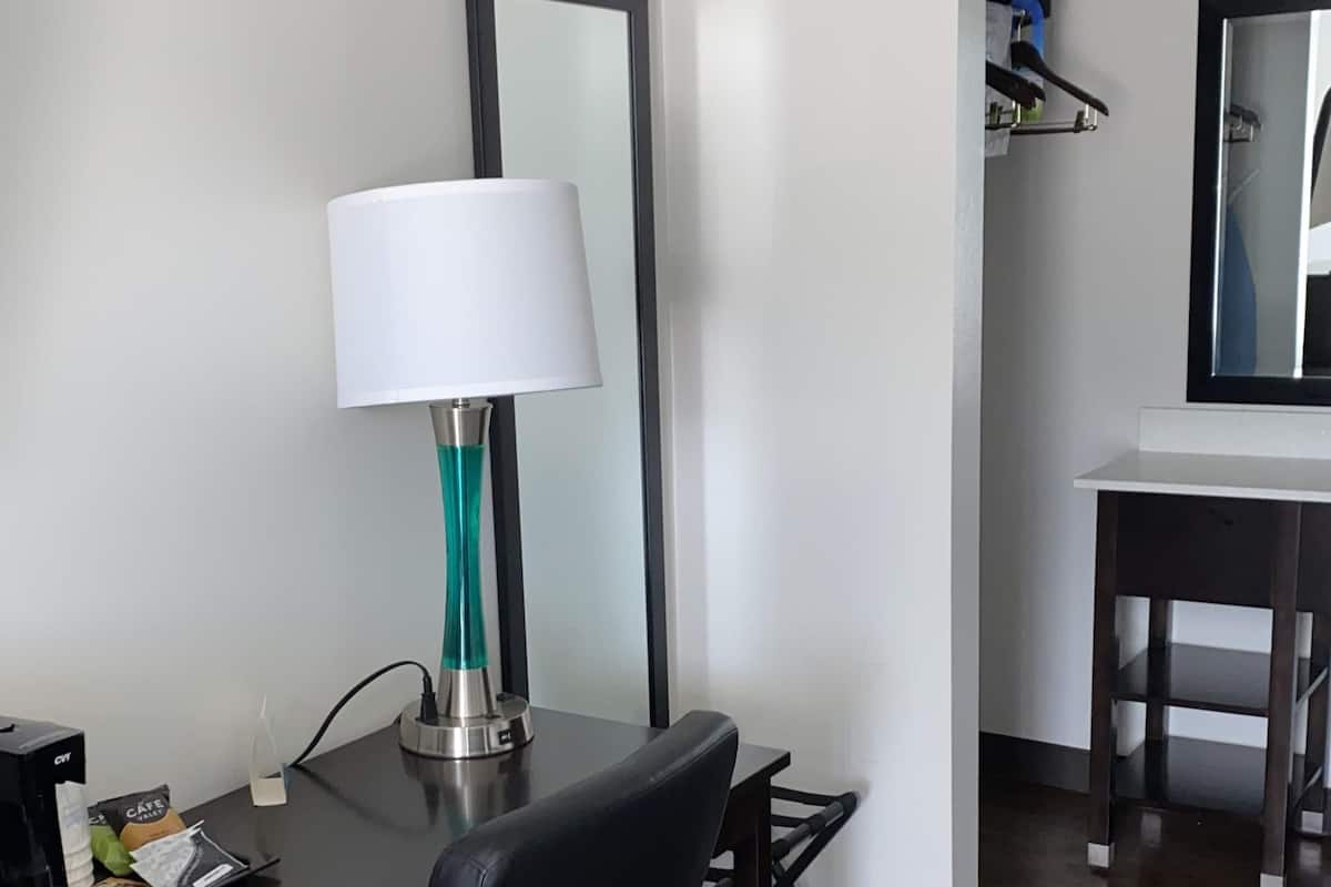 A hotel room with a desk, a lamp, a mirror, and a small table with a coffee maker and a bag of snacks.