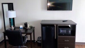 A hotel room with a desk, a chair, a lamp, a coffee maker, a microwave, a trash bin, and a TV mounted on the wall.