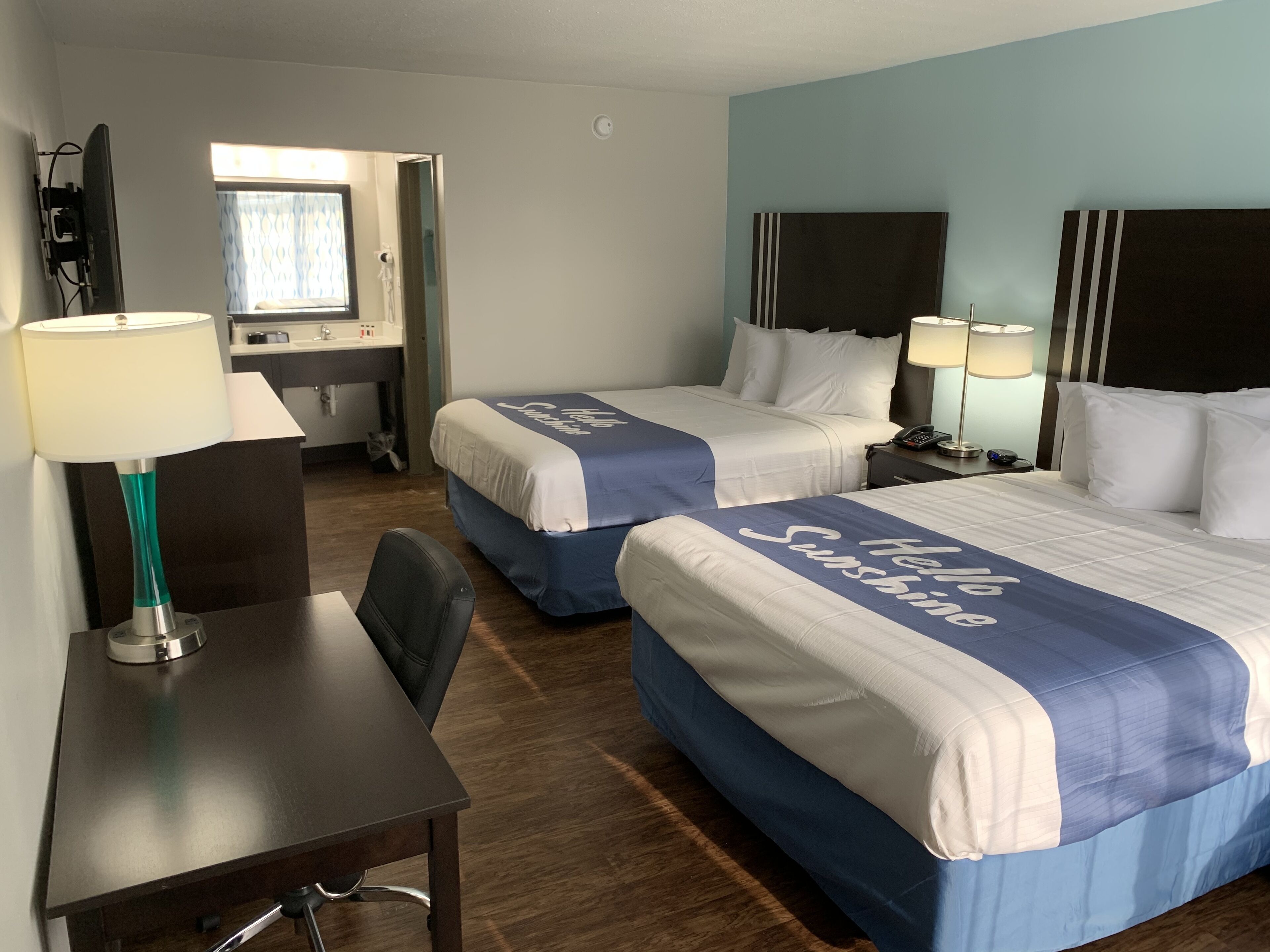 A hotel room with two beds, each with a 'Hello Sunshine' bedspread, a desk with a lamp, a chair, and a small table.