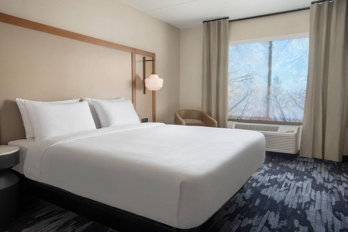 A hotel room with a large bed, a bedside table, a chair, a window with curtains, and an air conditioning unit.