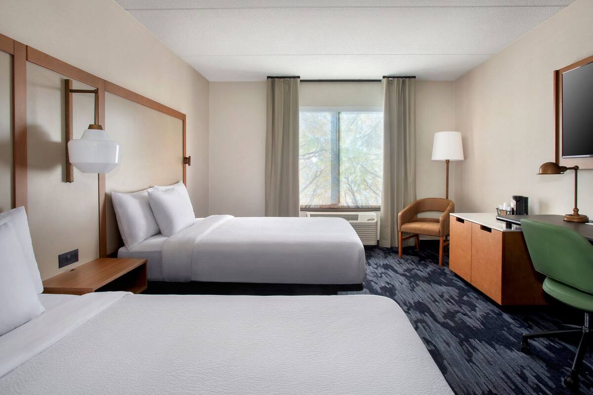 A hotel room with two beds, a desk, a chair, a lamp, and a large window with curtains.