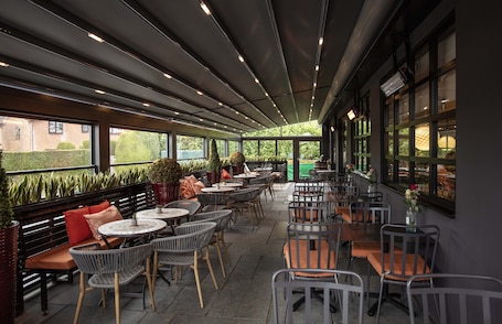 An outdoor cafe area with multiple tables and chairs, potted plants, and a view of the surrounding greenery.