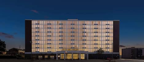 A modern hotel with a large glass facade, a central entrance, and a parking lot.